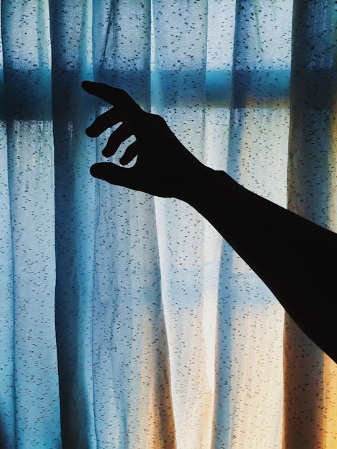 Silhouetted hand reaches toward softly lit curtain, creating a dramatic and moody scene indoors.
