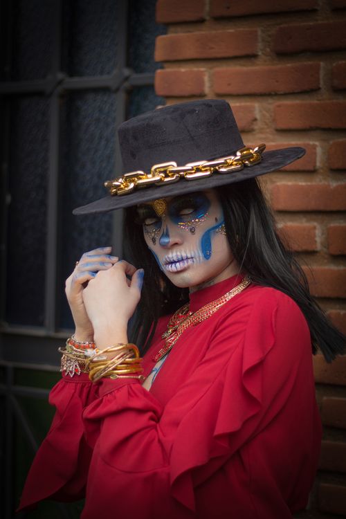 Free A woman with intricate Dia de los Muertos makeup poses in Mexico City, wearing traditional red attire and a chain-adorned hat. Stock Photo