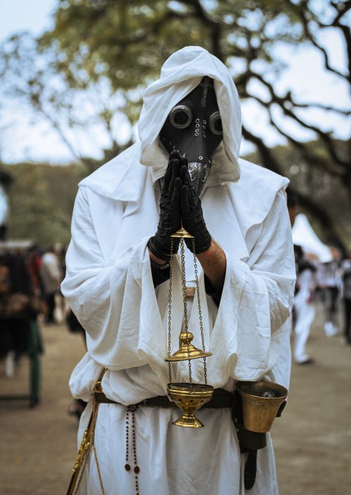 Free Hooded figure in white robe wearing mask, holding scales at outdoor festival. Stock Photo