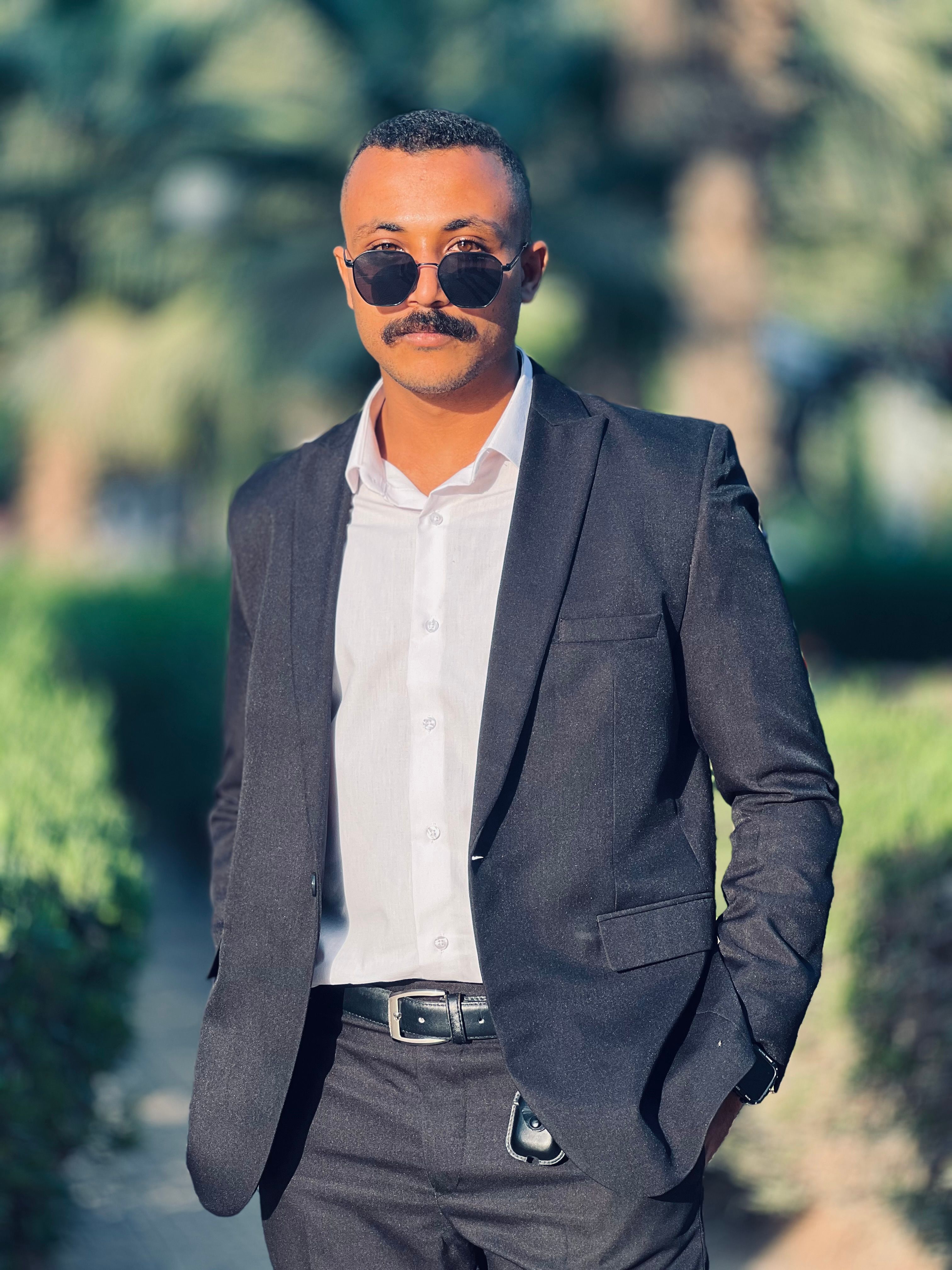 Confident adult in stylish suit and sunglasses posing outdoors with lush greenery backdrop.