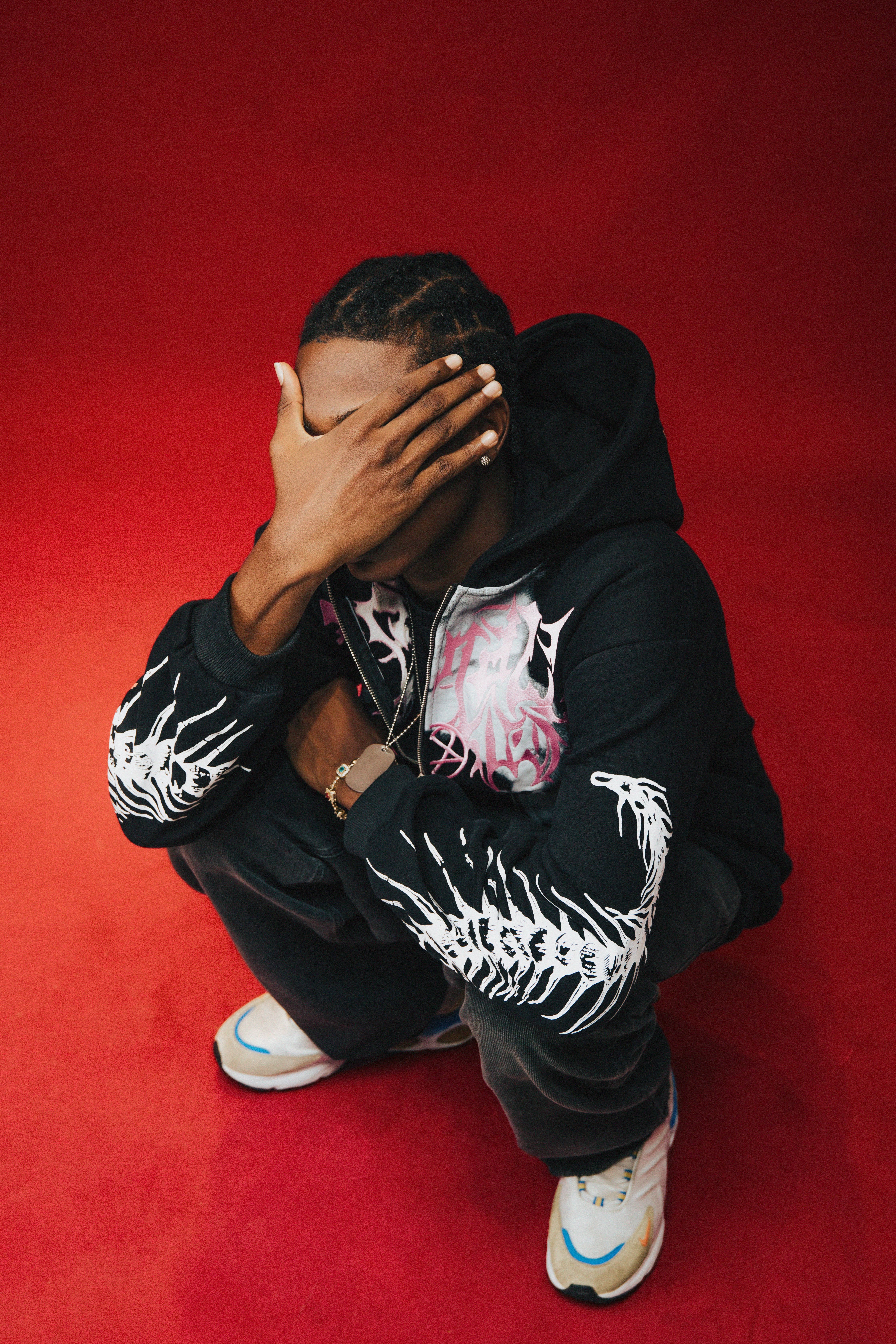 Fashionable young man in trendy streetwear squatting with hand on face against a red backdrop.
