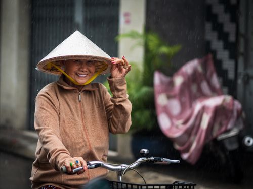 Free A smiling woman rides a bicycle in Ho Chi Minh City, wearing a conical hat during a gentle rain. Stock Photo