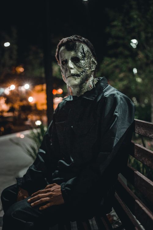 Free A mysterious figure wearing a mask sits on a park bench at night, creating an eerie urban atmosphere. Stock Photo