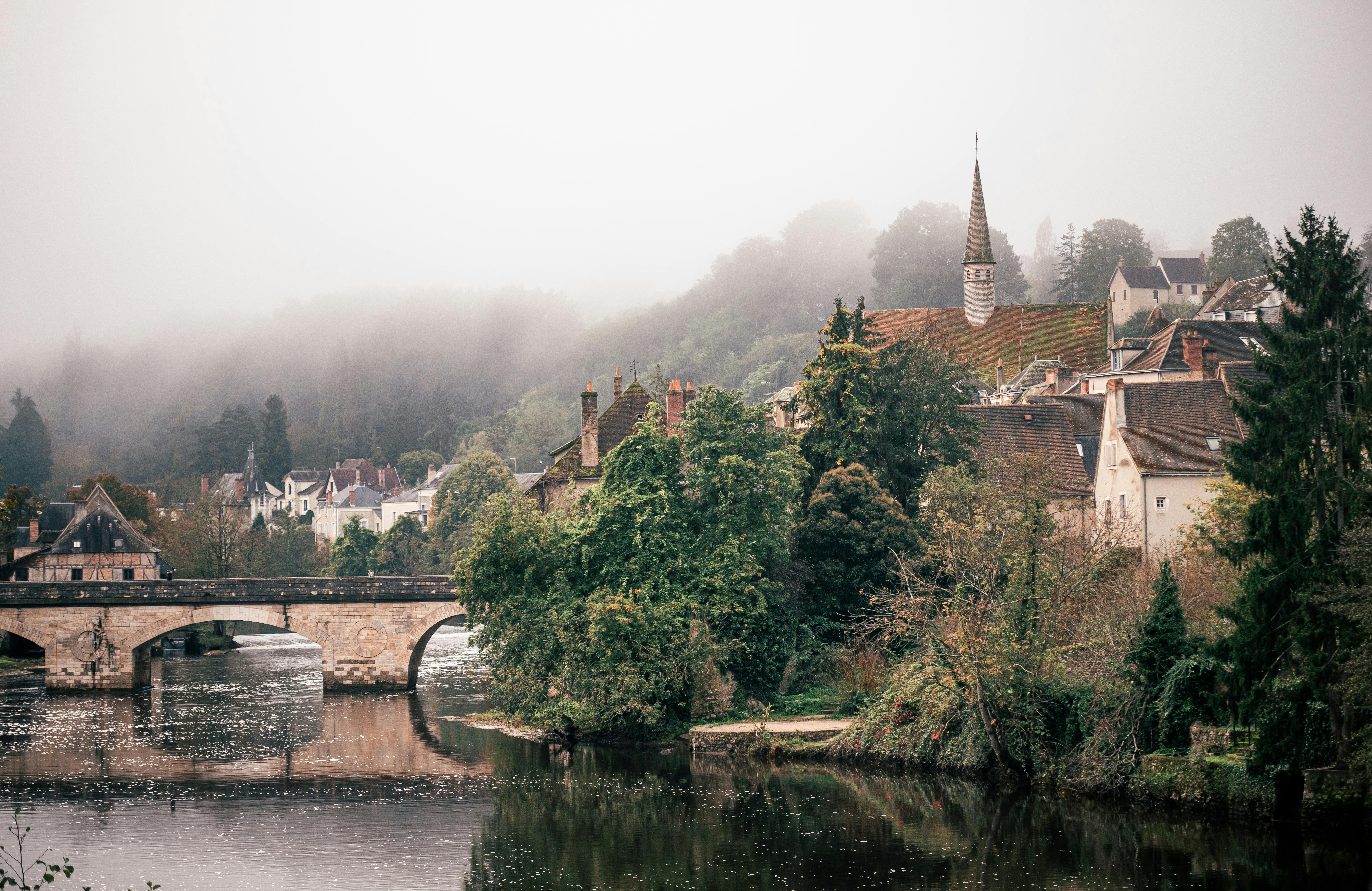 Charming misty morning view of a French village with a historic stone bridge over a river.