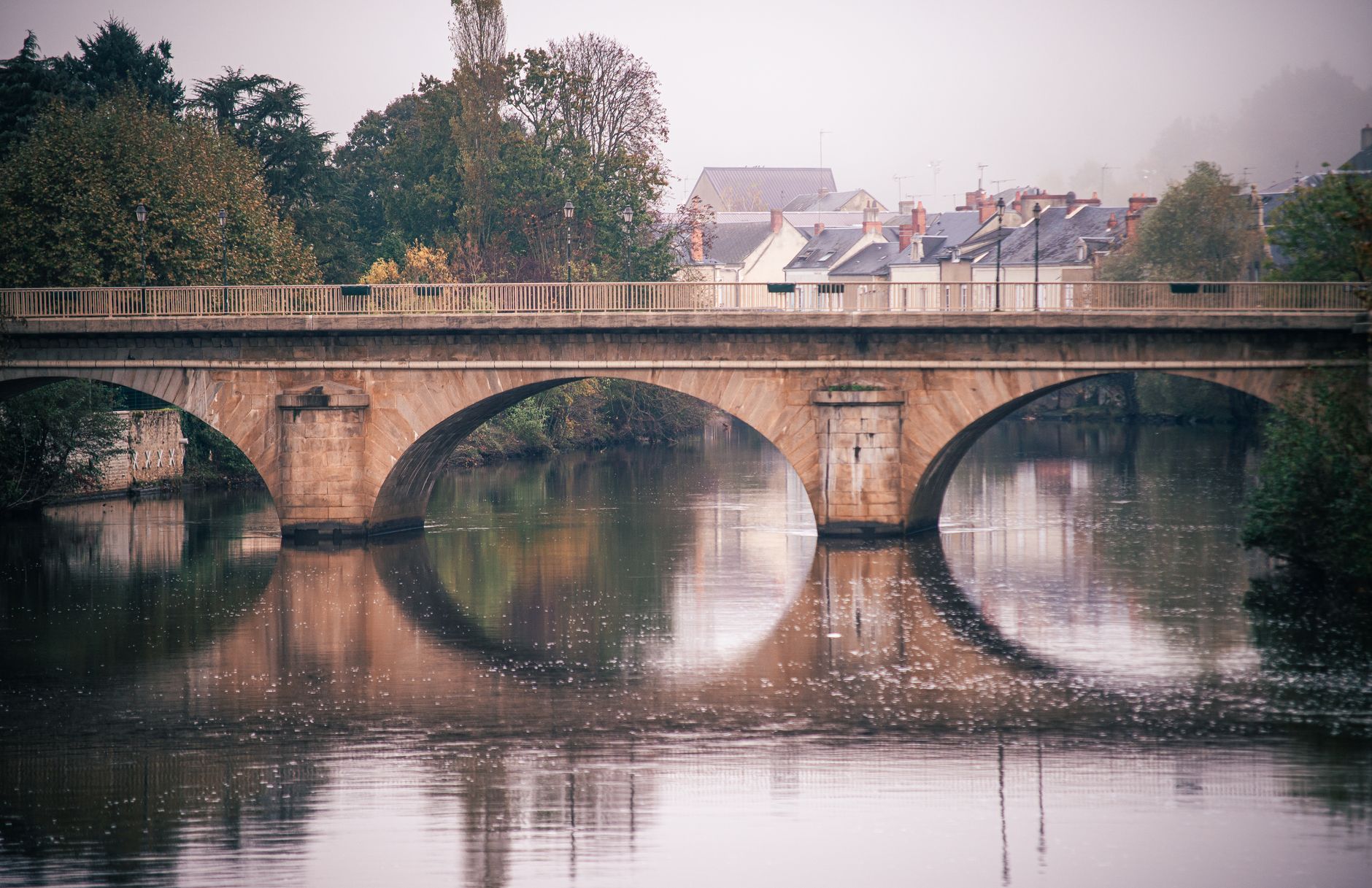 A scenic view of a stone bridge over a calm river with misty houses in the background.