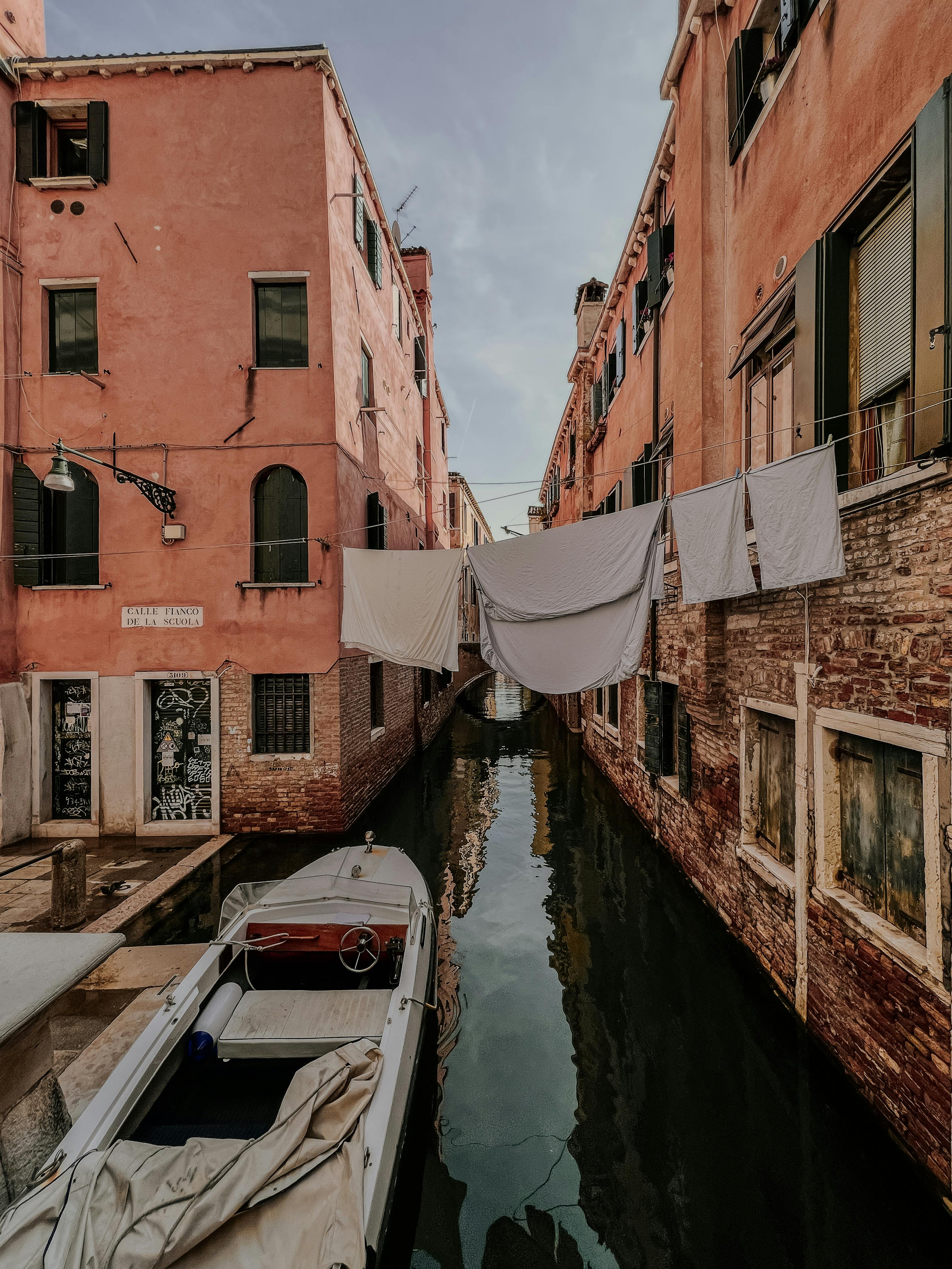 A narrow Venetian canal with boats and laundry hanging above, framed by pink buildings.
