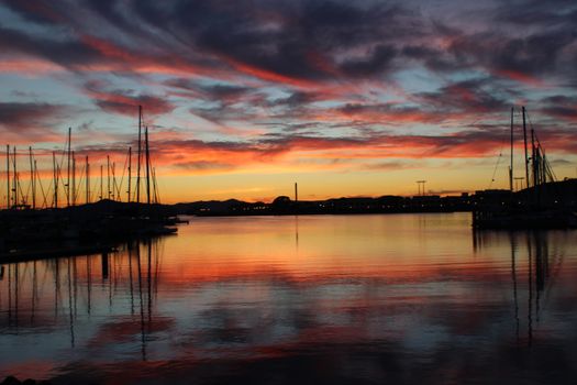 A stunning sunset over a harbor with sailboats, reflecting vibrant sky colors on the water.