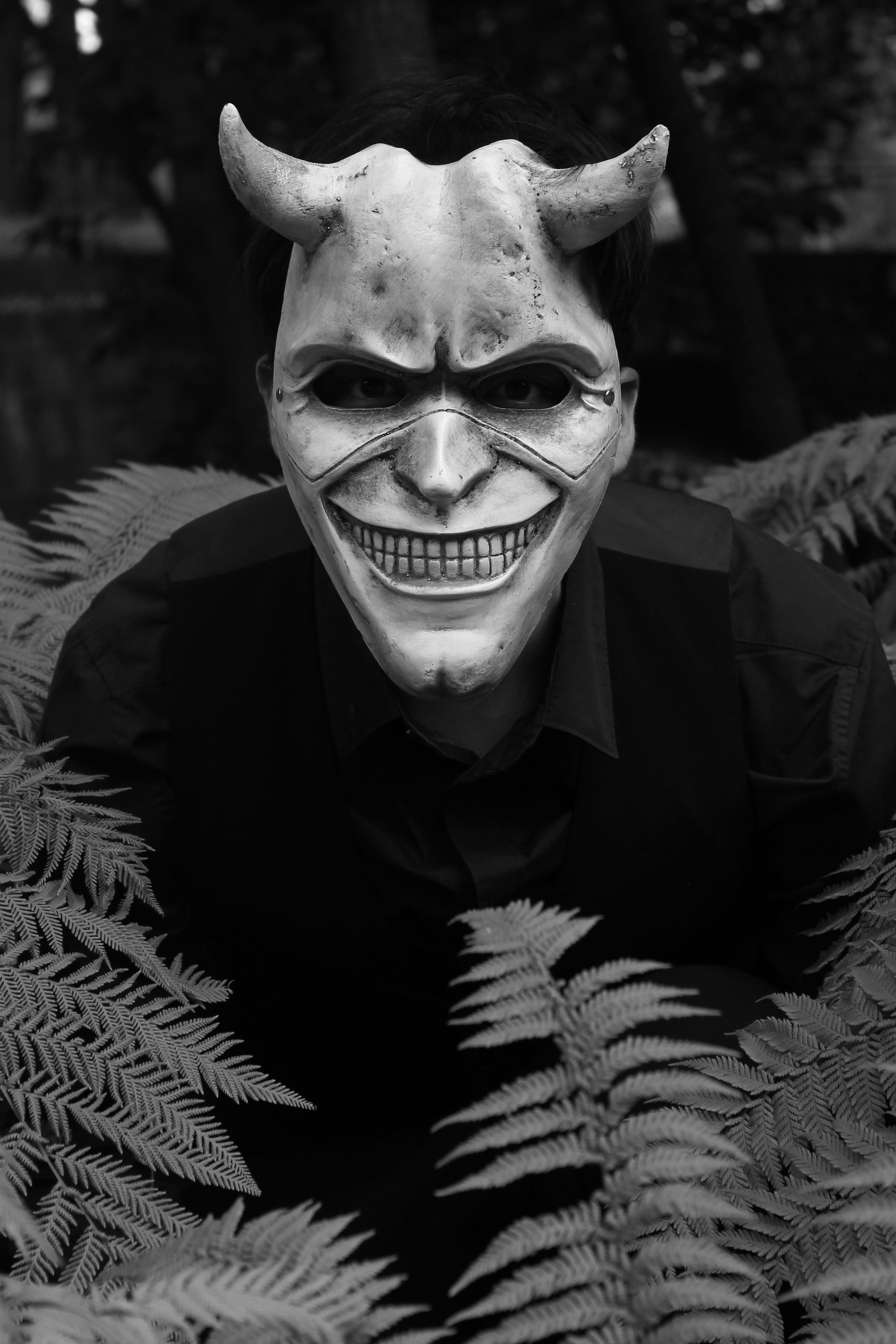 A man wearing a devil mask crouches among ferns in a dark outdoor setting.