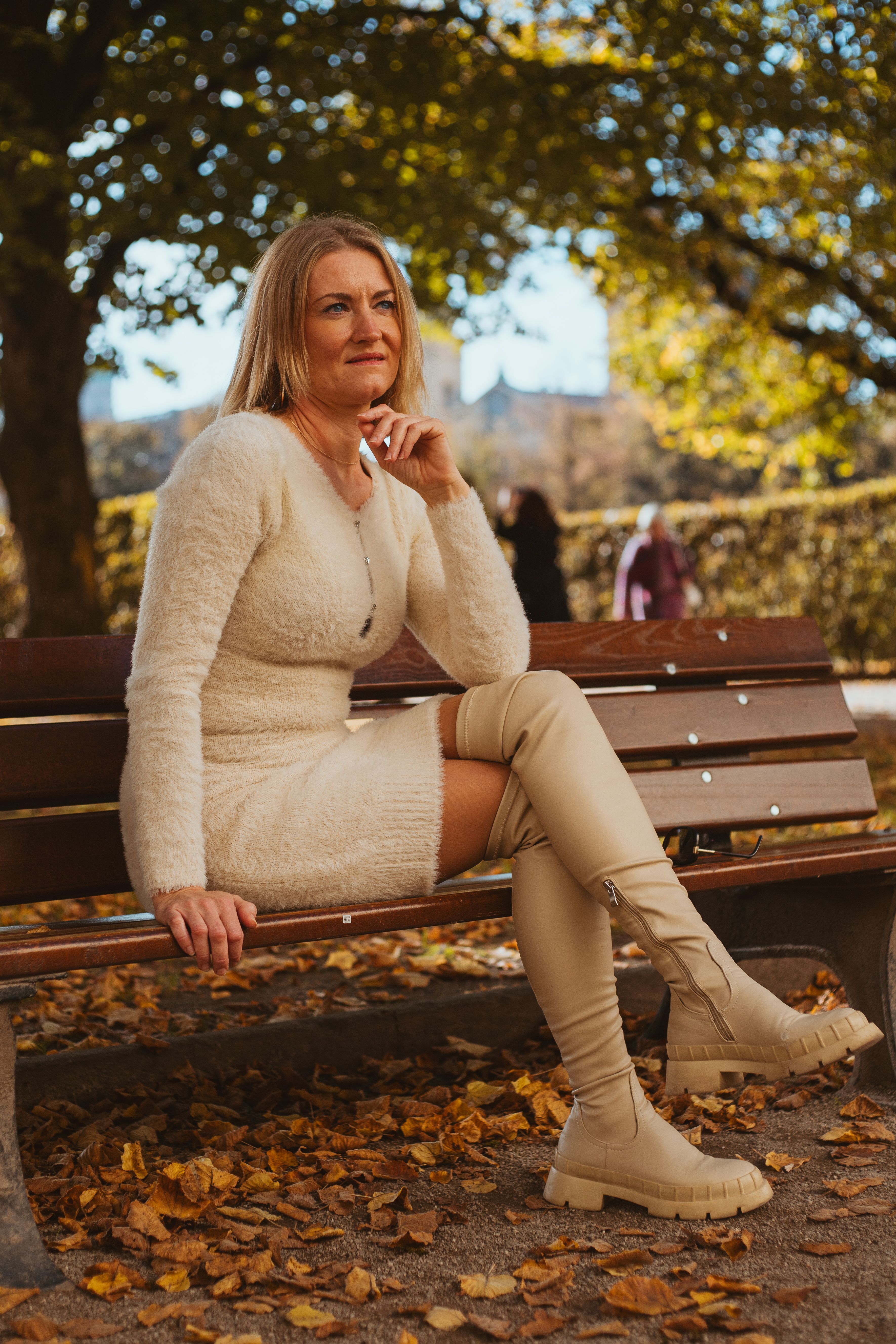 A stylish woman sits gracefully on a bench in a Munich park surrounded by autumn leaves.