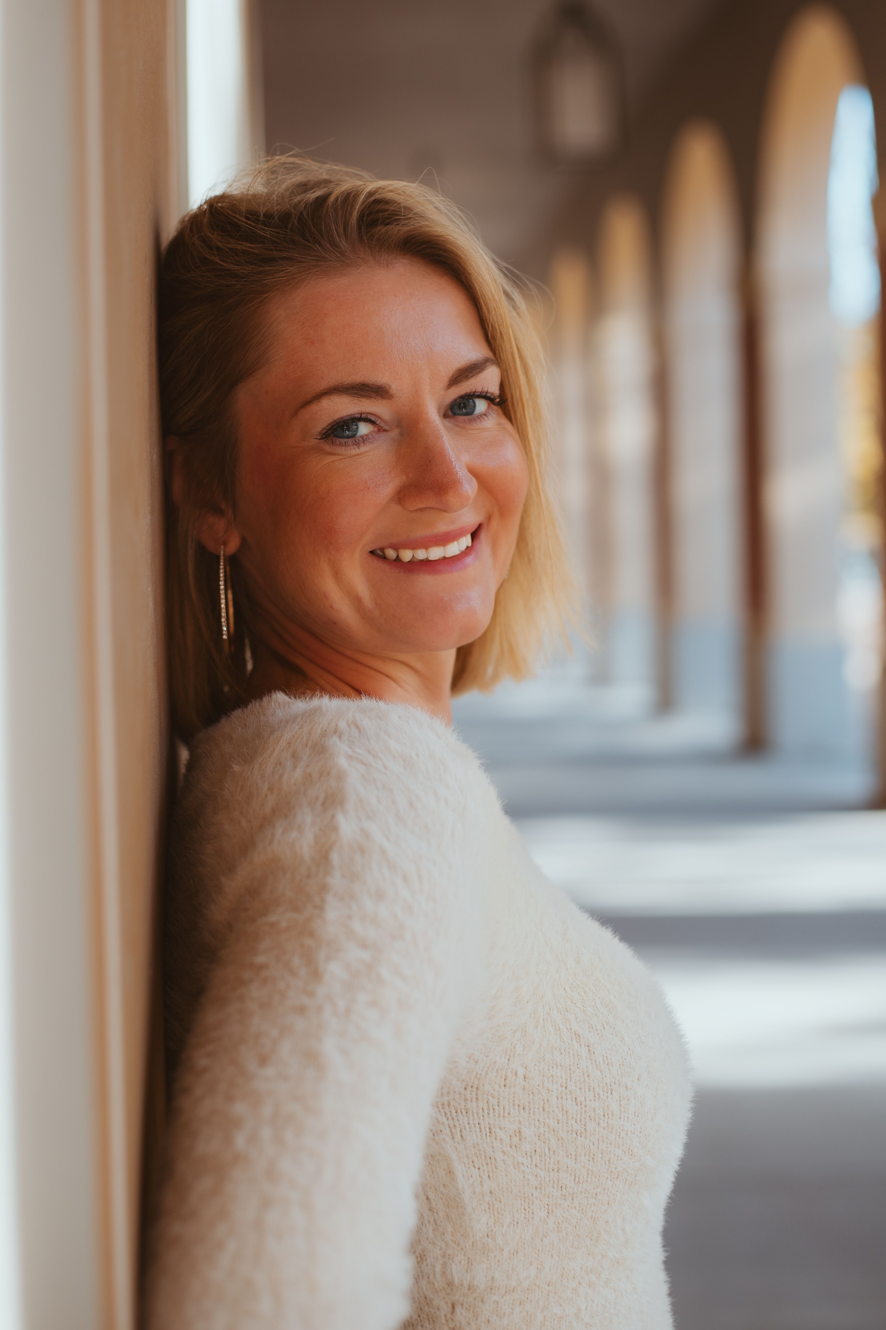 Cheerful woman leaning against a wall in Munich's architectural setting, bathed in sunlight.