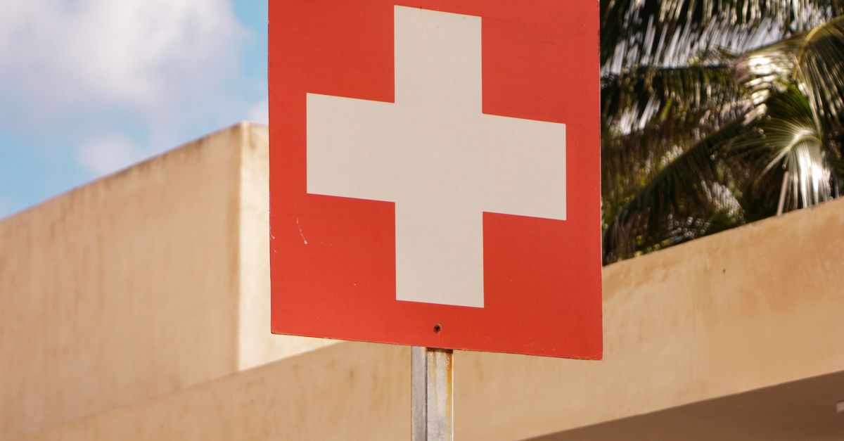 Photo by Rodrigo Ortega A red healthcare cross sign stands in front of a building with palm trees, under a bright blue sky.