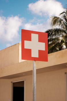Photo by Rodrigo Ortega A red healthcare cross sign stands in front of a building with palm trees, under a bright blue sky.