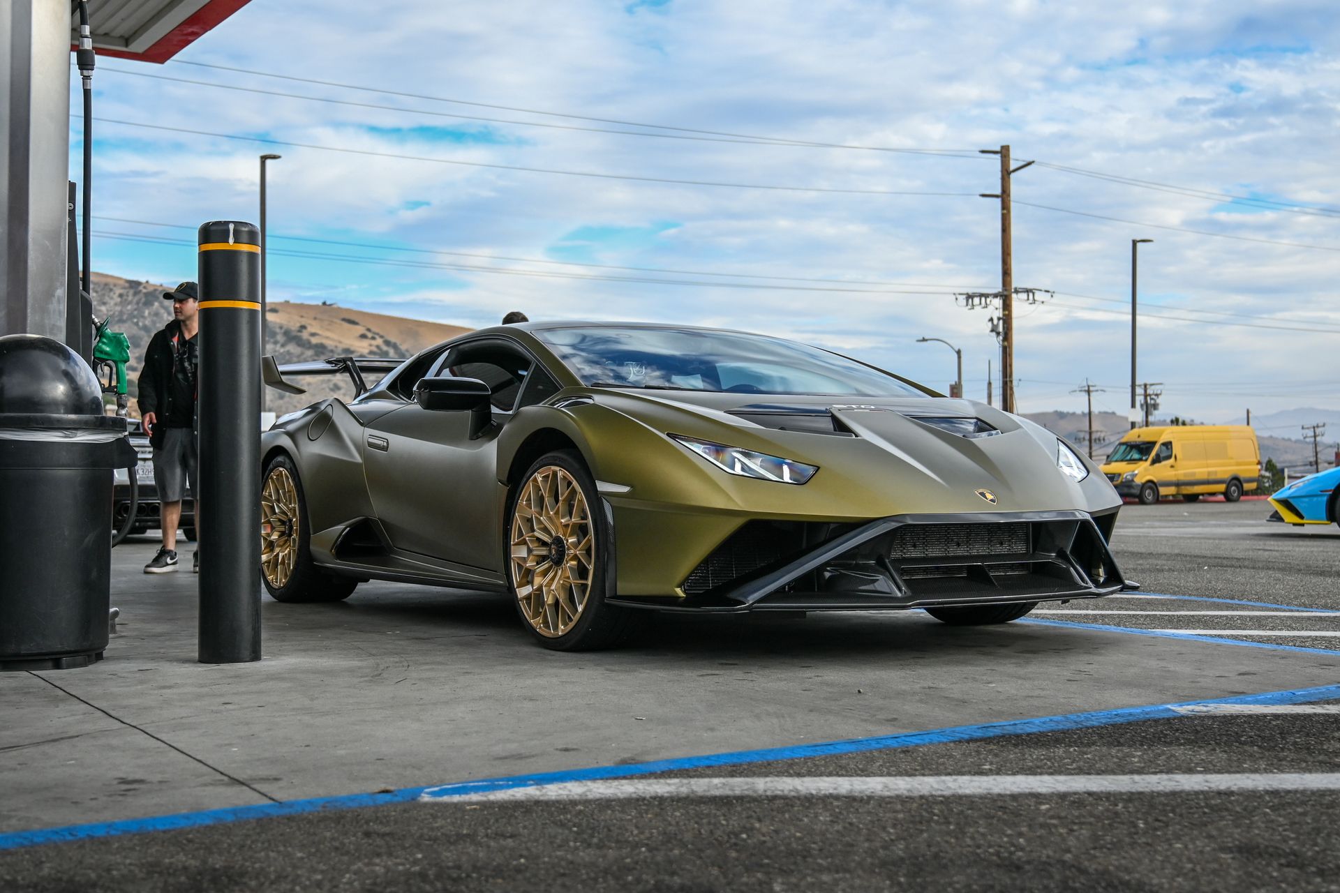 Luxury olive green sports car parked at a gas station under a bright sky.