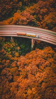 An aerial shot of a truck driving on a curved road surrounded by vibrant autumn foliage.