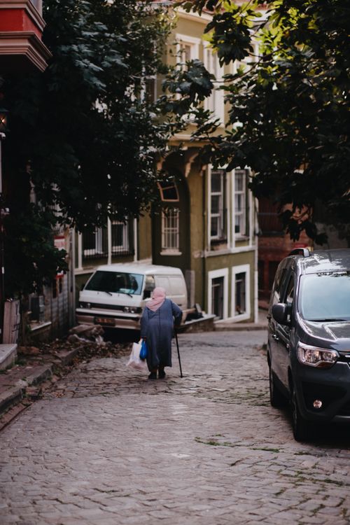 Free An elderly woman walks on a quaint cobblestone street lined with parked cars and old buildings. Stock Photo