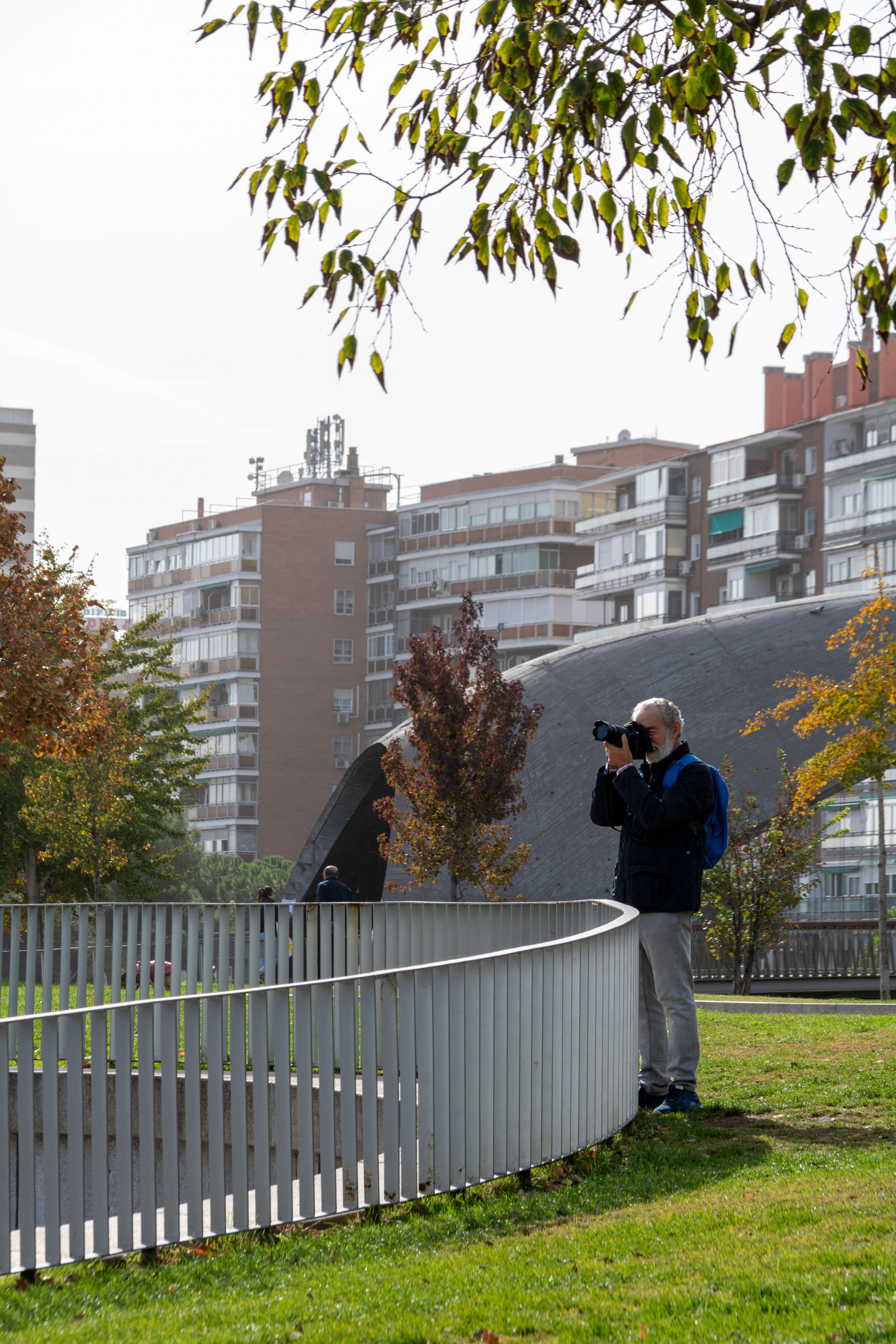 A photographer in a Madrid park capturing autumn city life with residential buildings in the background.