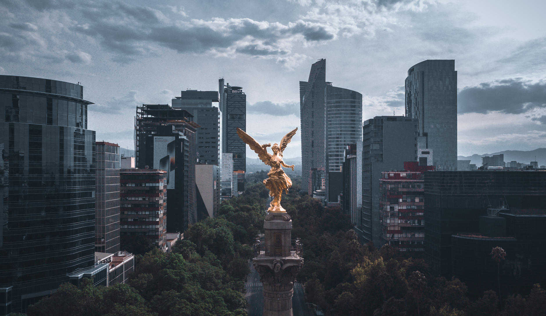 Aerial view of the Angel of Independence with Mexico City skyline, showcasing modern buildings and dramatic sky.