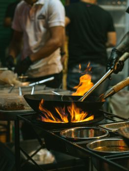 An energetic scene of Asian street food being cooked in a wok with flames on a bustling street.