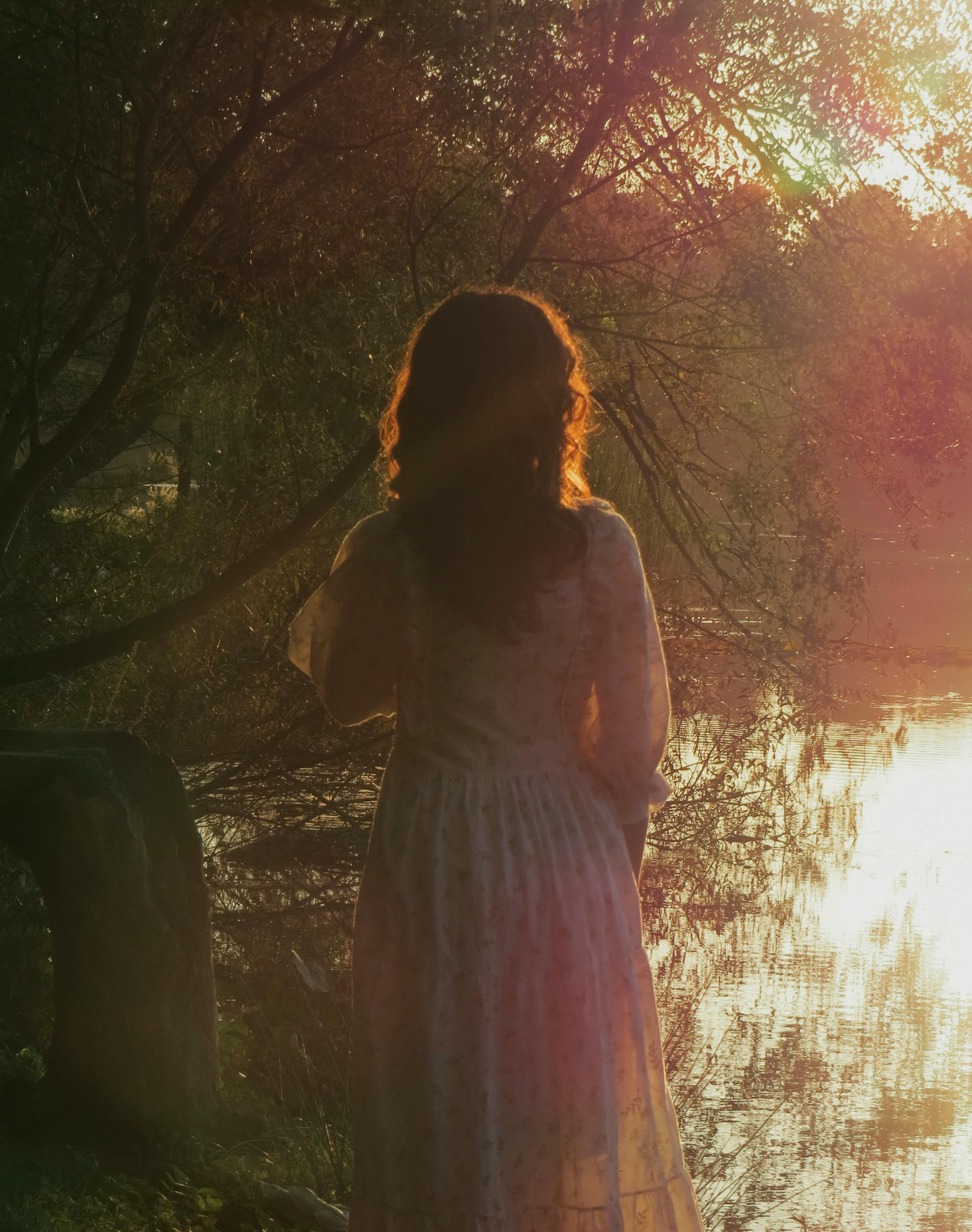 A woman in a vintage dress stands by a calm lake at sunset, surrounded by nature.