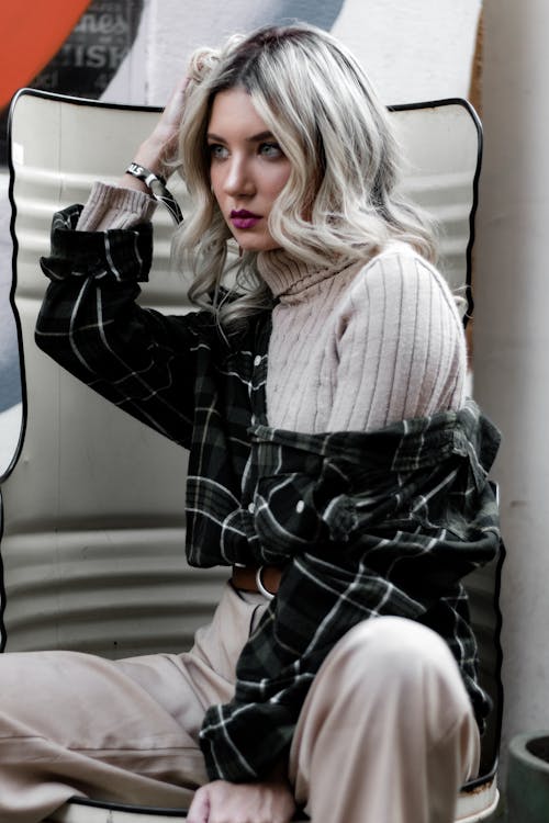 Confident blonde in sweater and shirt sitting on chair and looking away while touching hair