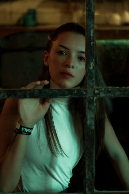 Young contemplative female in trendy wear looking at camera behind old window at home in evening