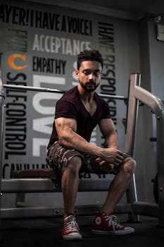 Male fitness model sitting on gym equipment, showcasing muscles and strength.