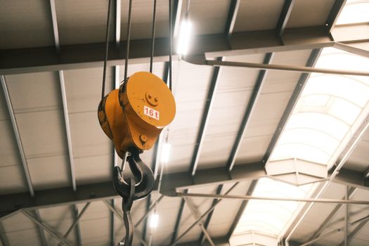 Closeup of a yellow crane hook hanging from ceiling in a large industrial warehouse.