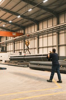 A worker operates an overhead crane in an industrial warehouse in Konya, Türkiye, lifting metal pipes.