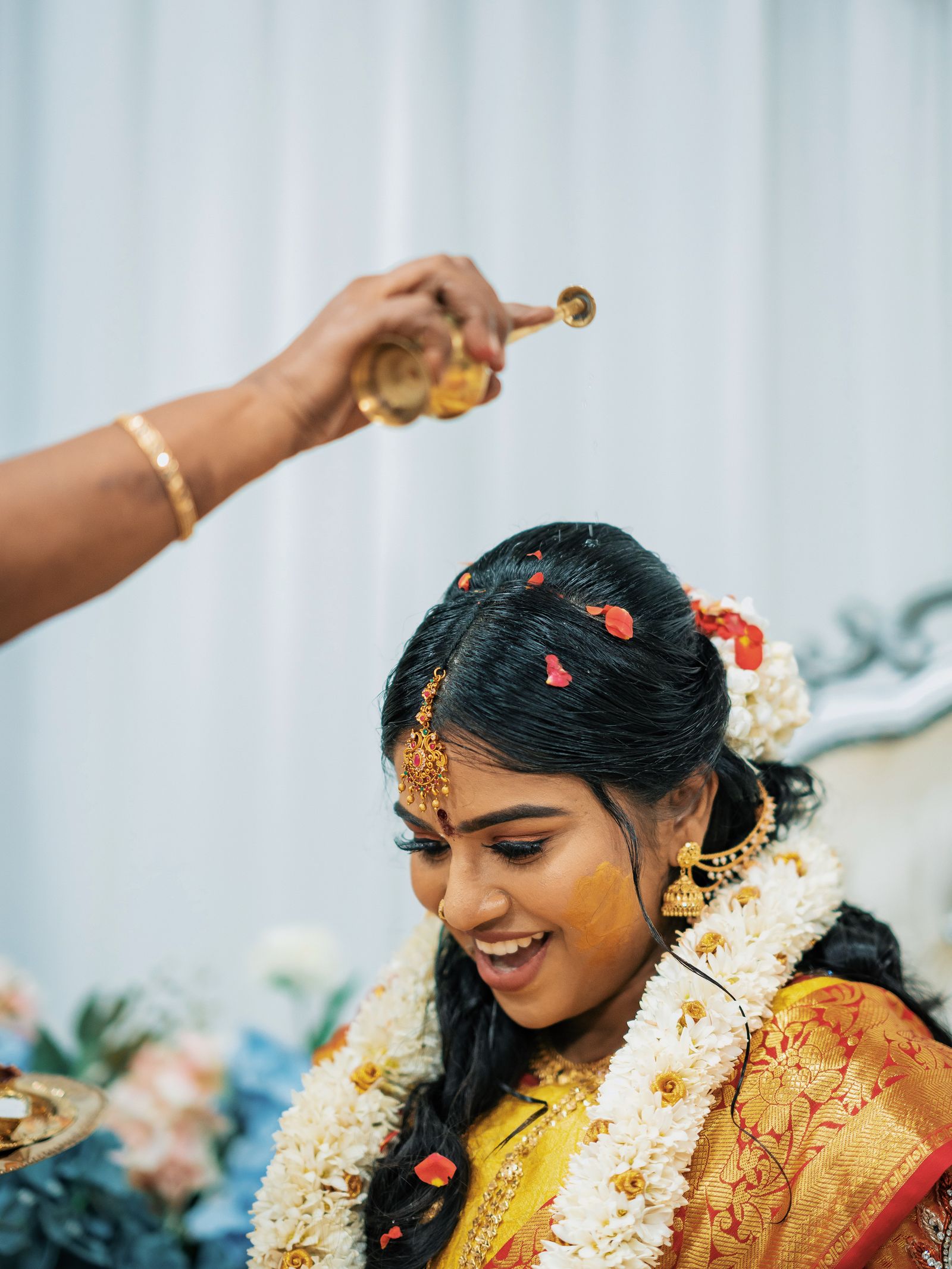 A bride laughs with her eyes closed as an elder pours from a small gold kalash above her head, turmeric on her cheek and rose petals in her hair.