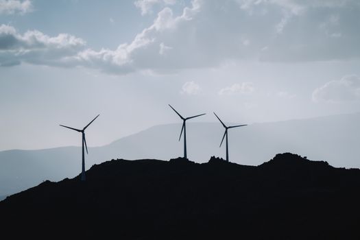 Silhouette of three wind turbines on a mountain ridge under a cloudy sky, promoting renewable energy.