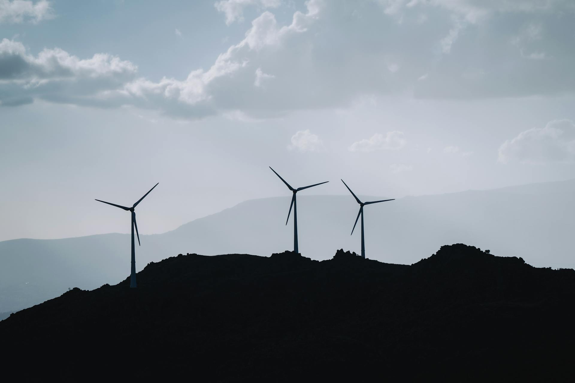 Silhouette of three wind turbines on a mountain ridge under a cloudy sky, promoting renewable energy.