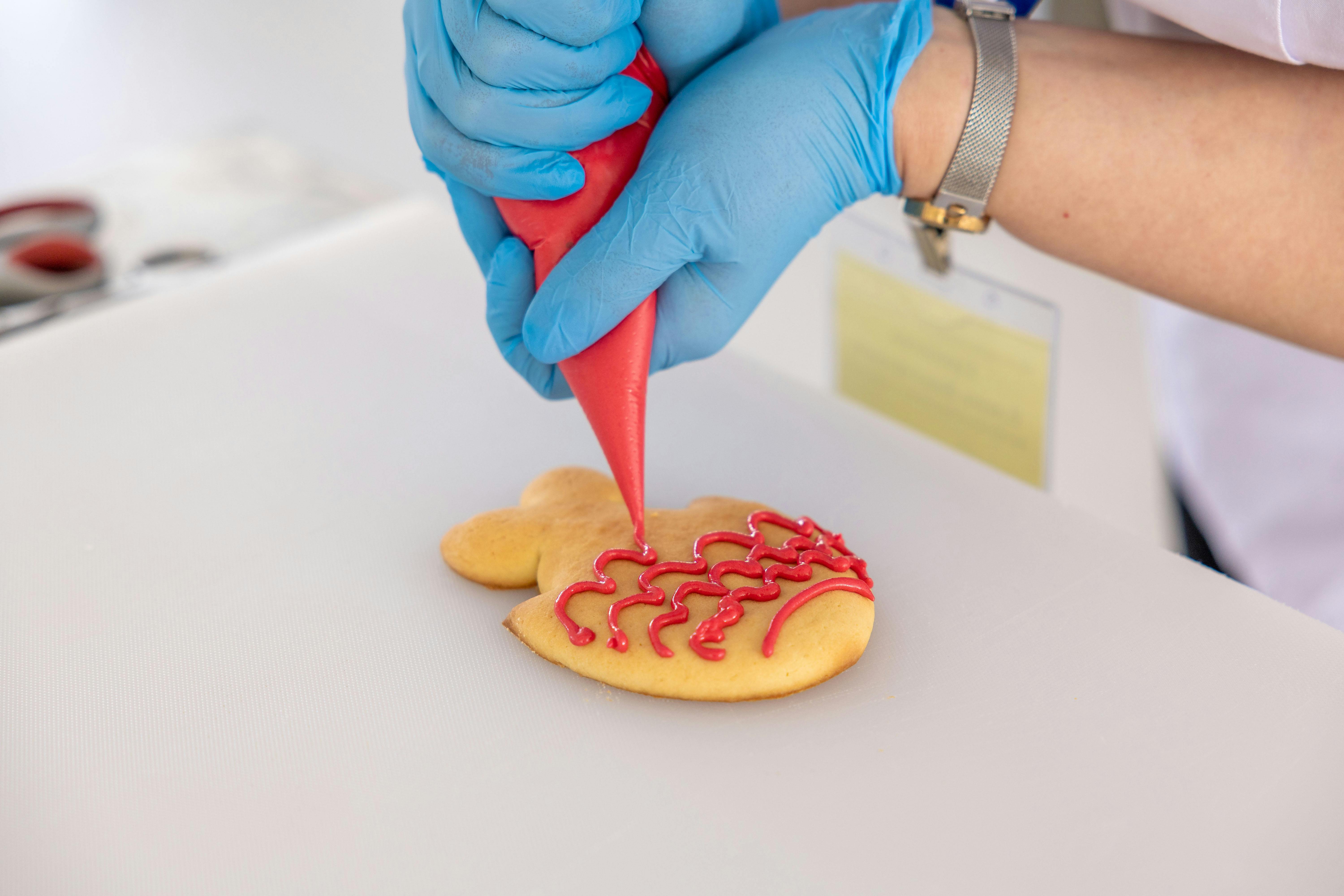 Close-up of a cookie being decorated with red icing by a gloved hand.