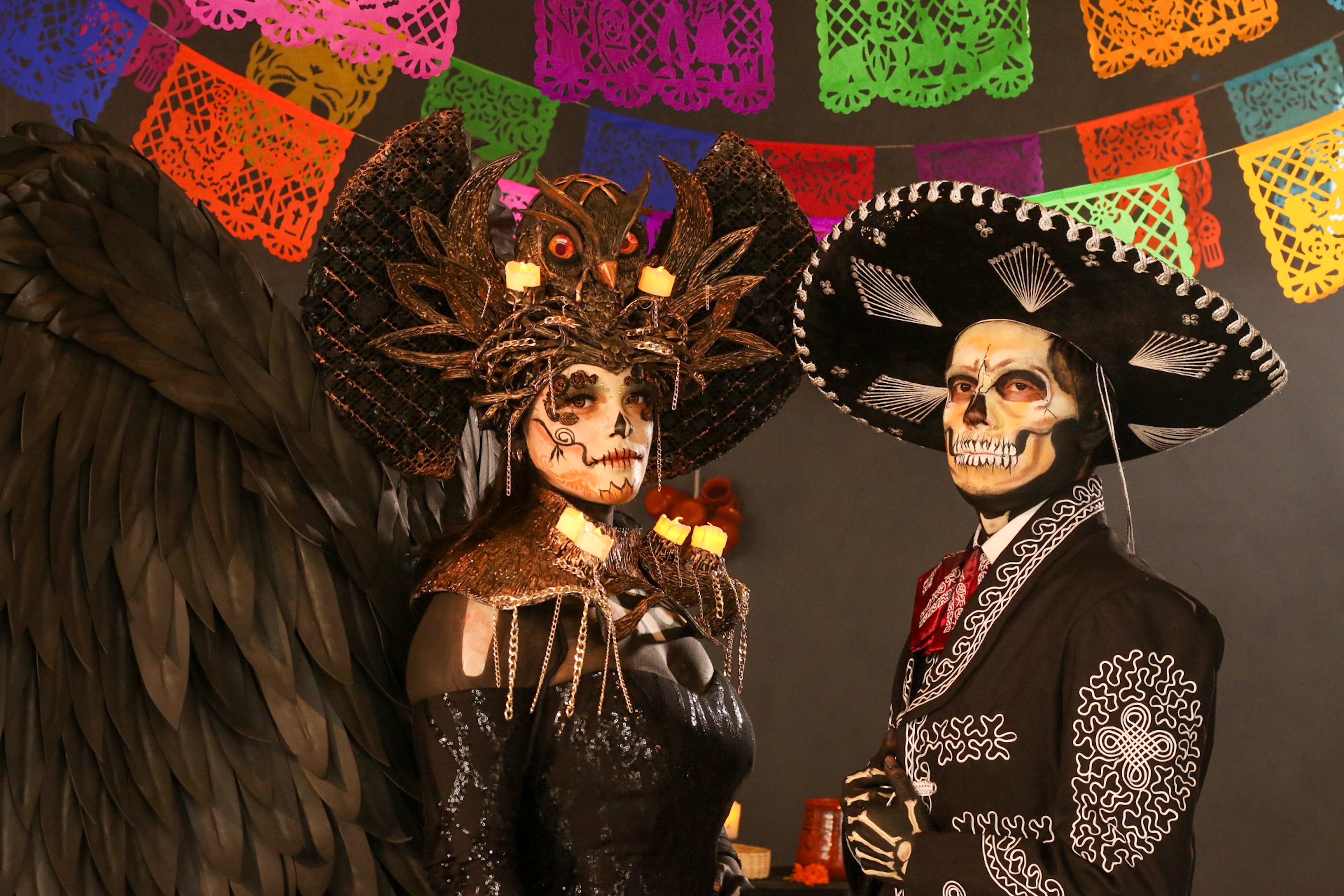 Two adults in ornate Day of the Dead costumes with colorful papel picado decor.