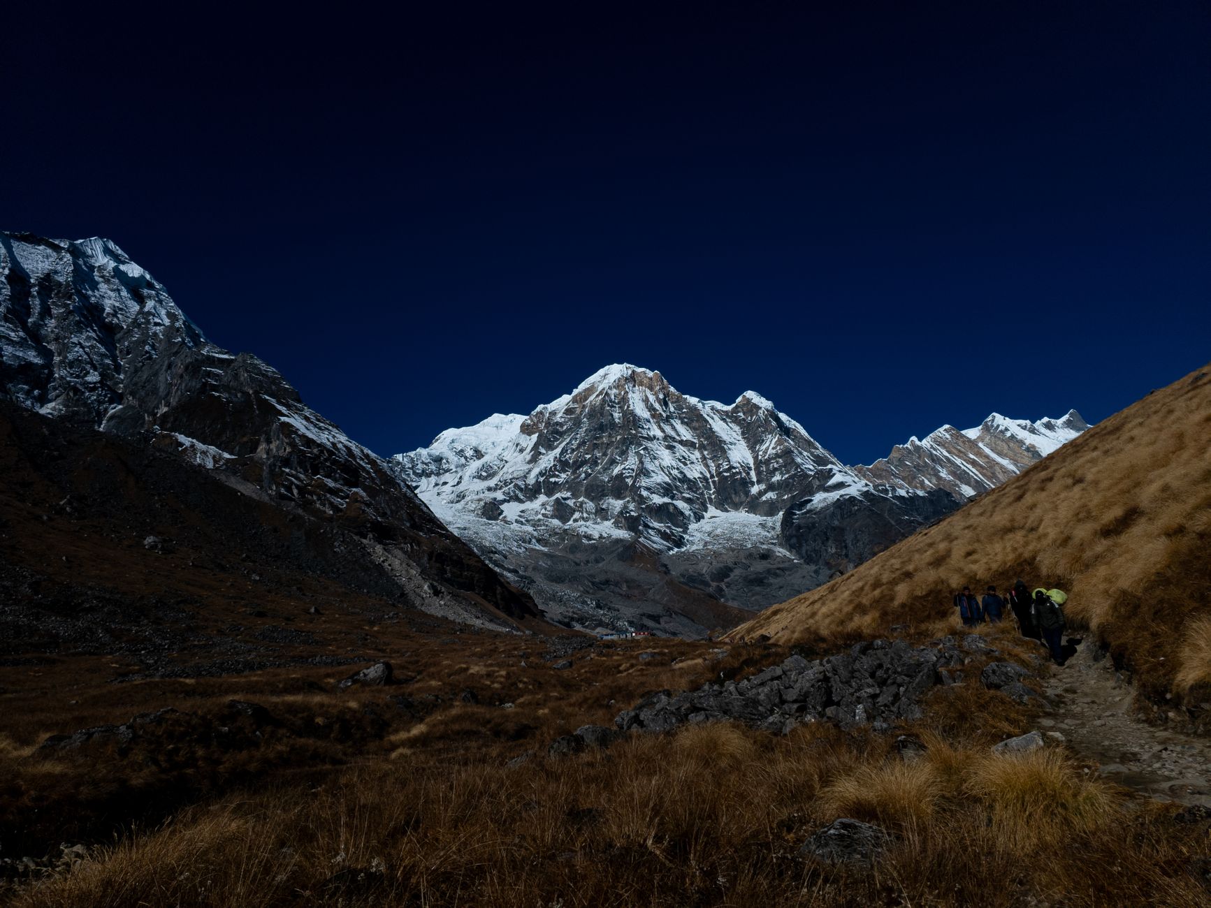Trekkers in the Himalayas ascend towards a breathtaking snow-capped peak under a vibrant blue sky.