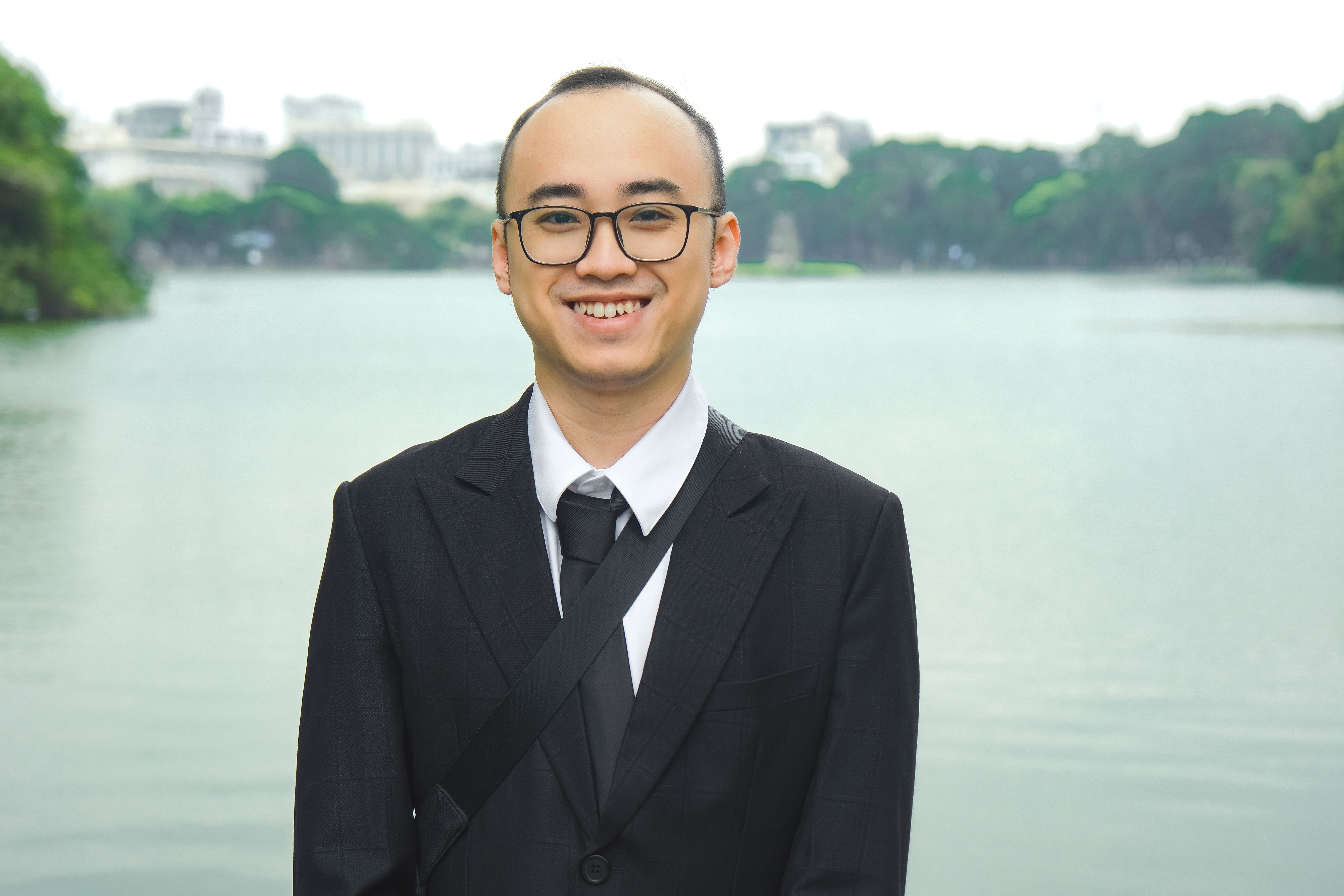 Young professional in a black suit smiling by a serene lakeside backdrop.