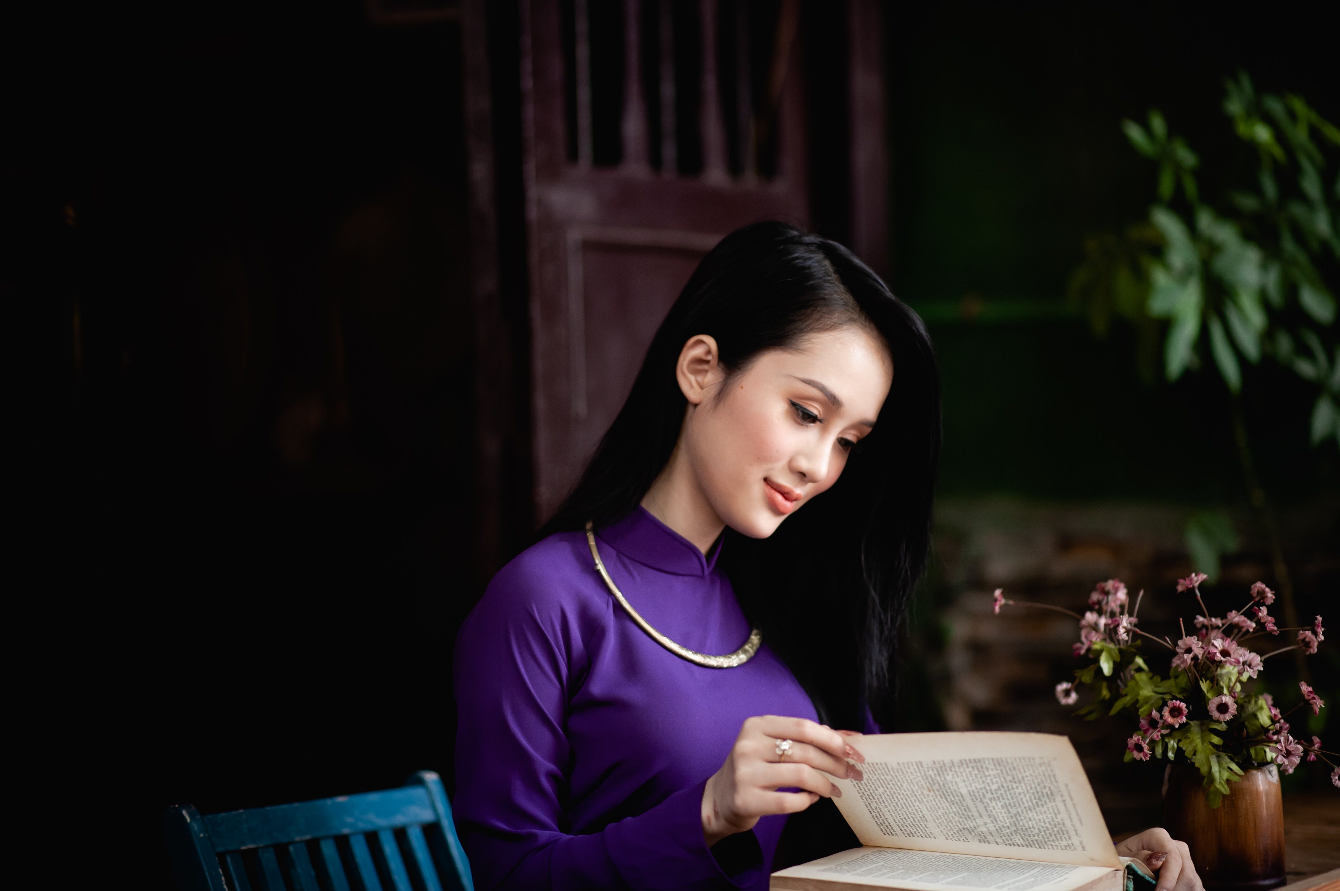 A Vietnamese woman wearing a purple Ao Dai reads a book, indoors surrounded by flowers.