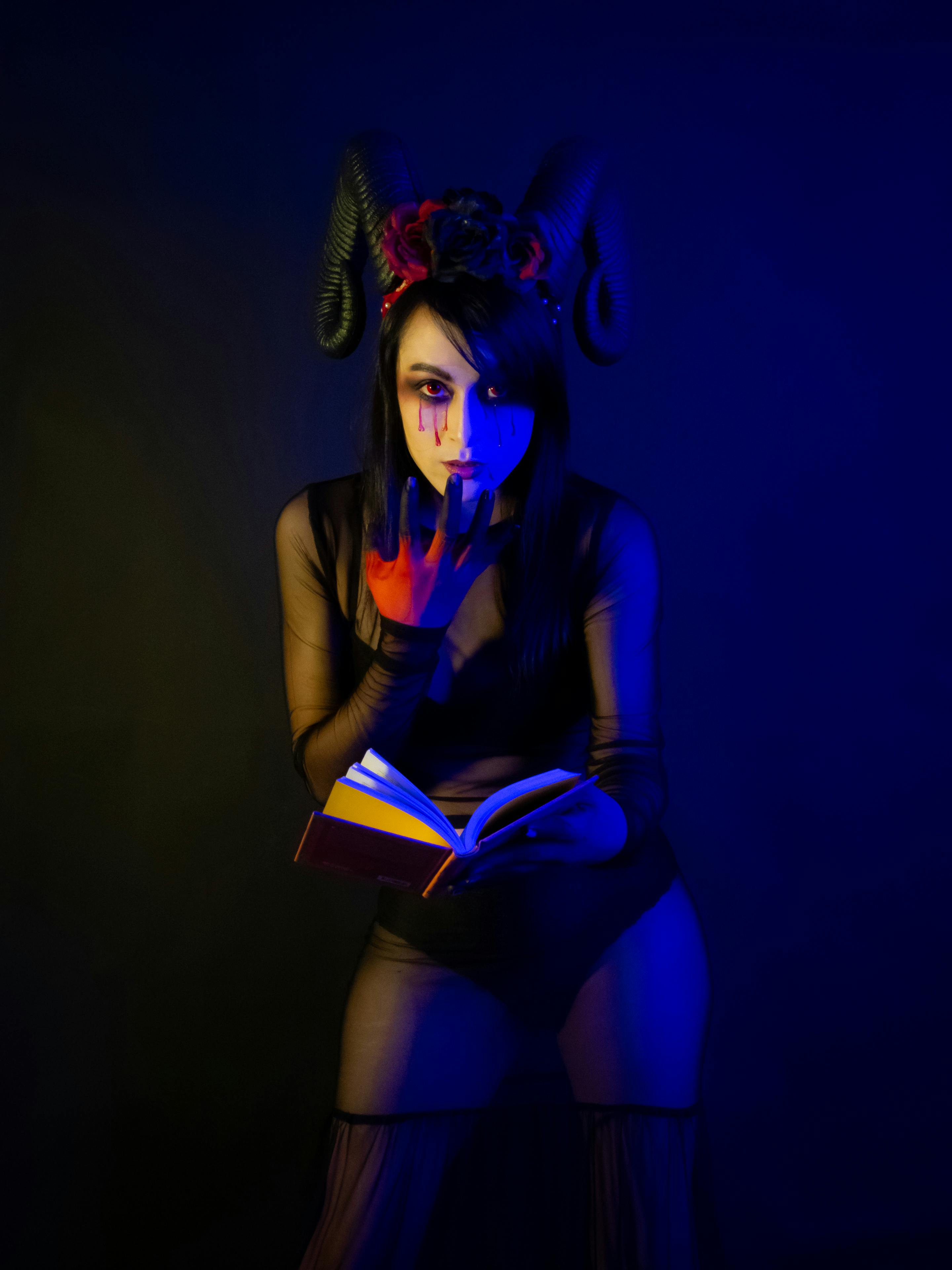 grátis Mulher em traje gótico com chifres, lendo um livro, iluminação ambiente escura. Foto profissional