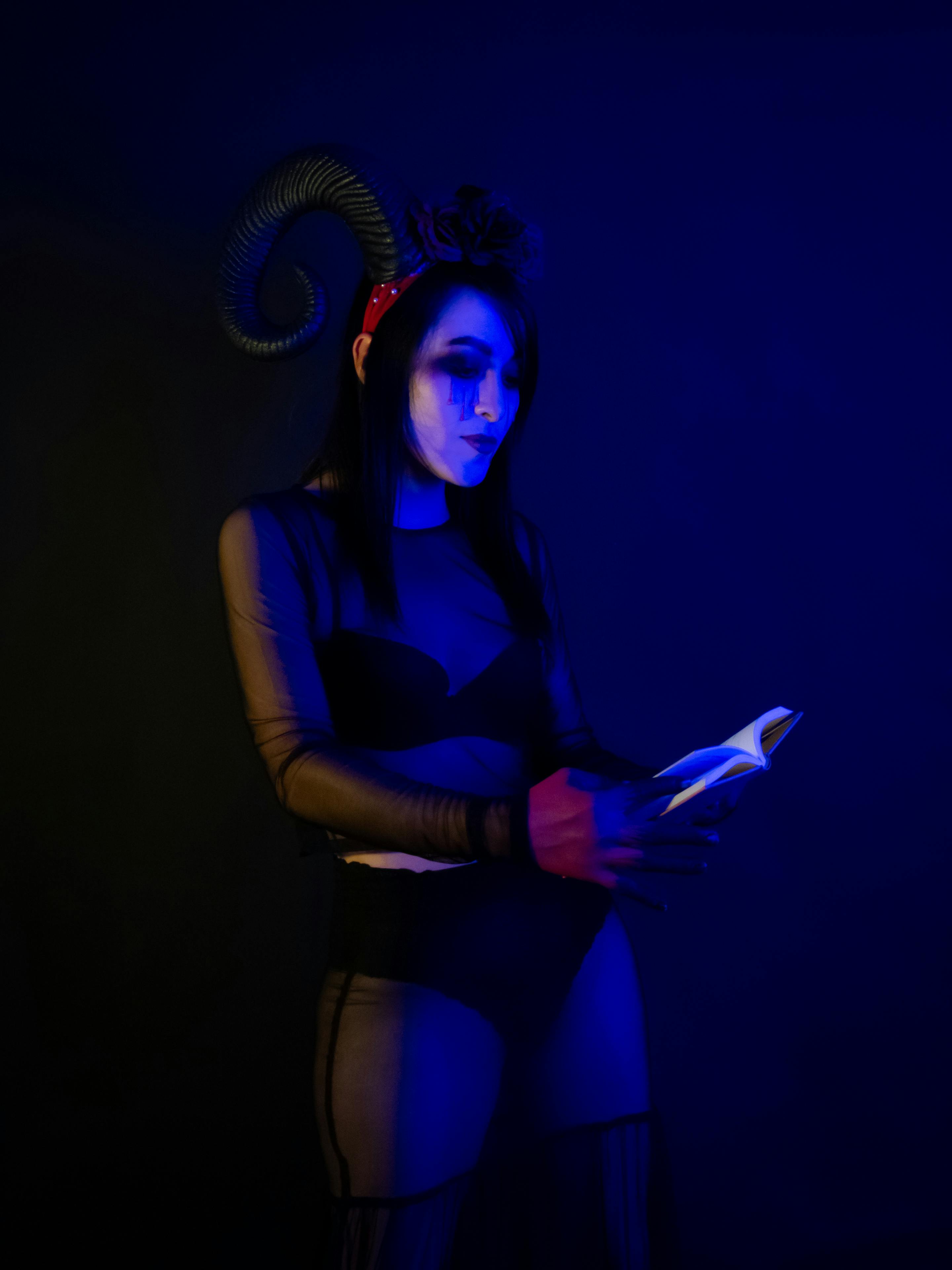 grátis Um artista fantasiado de diabo lê sob iluminação azul, capturando uma atmosfera de Halloween. Foto profissional