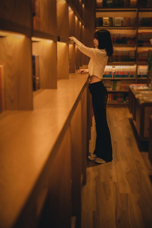 Free A young woman reaching for a book in a warmly lit library aisle. Stock Photo