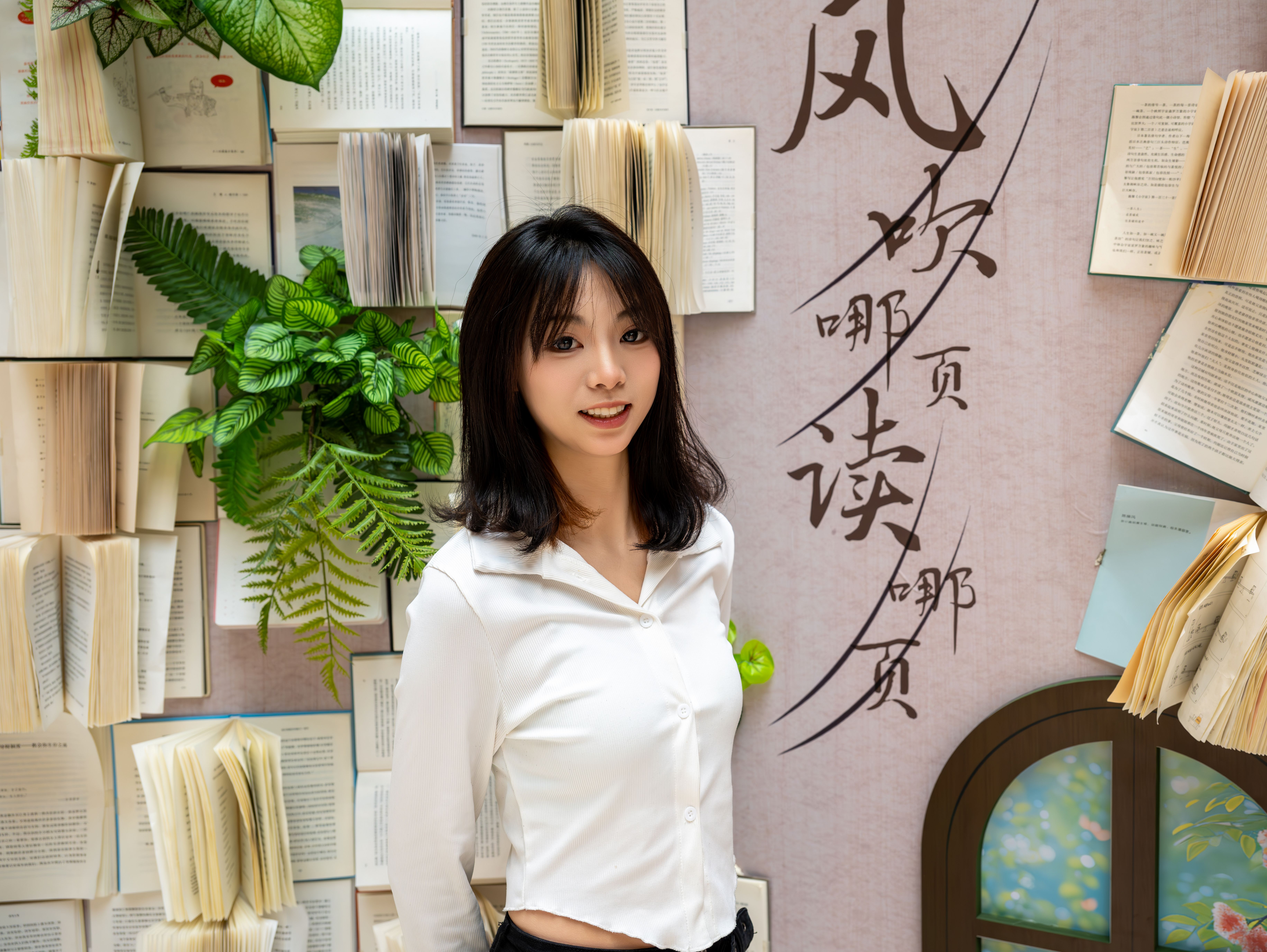 Portrait of a smiling woman with open books and plants on a decorative wall.
