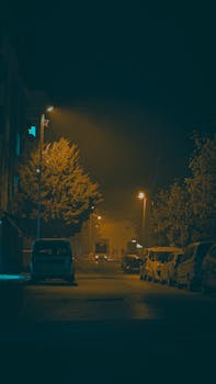 Dimly lit urban street at night with parked cars and streetlights casting a warm glow.