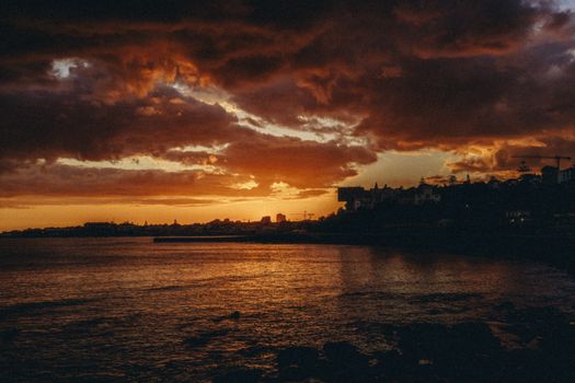 Stunning sunset over the coastline of Cascais, Portugal, with vibrant clouds and silhouetted buildings.