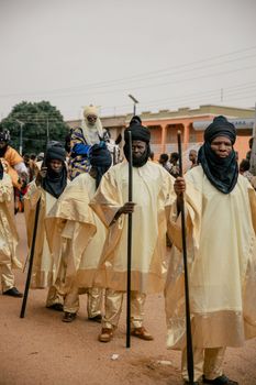 Men in traditional attire participate in an African cultural parade with vibrant ceremonial robes and walking sticks.
