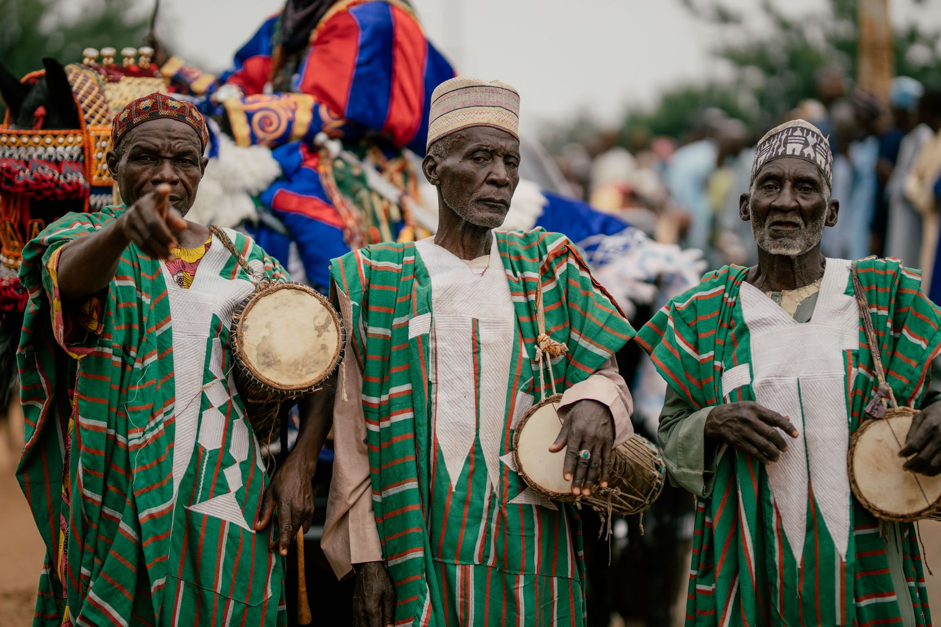 Three senior African men in traditional attire playing drums at an outdoor cultural event.