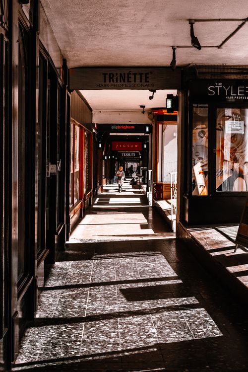 Free A sunlit urban hallway showcasing shops, shadows, and a walking individual. Stock Photo
