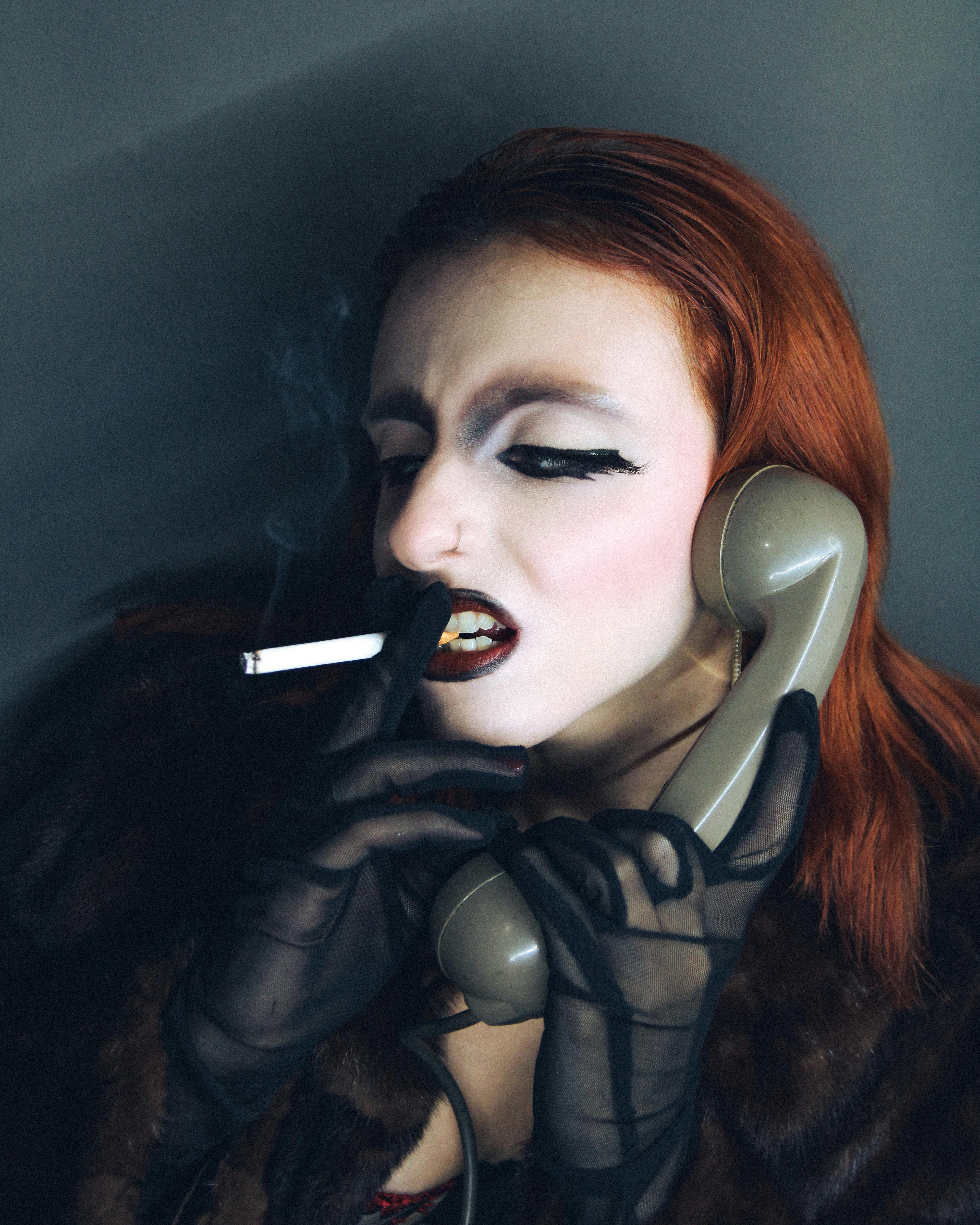 Gratis Ritratto intenso di una donna dai capelli rossi con un trucco teatrale, che tiene in mano un telefono d'epoca e una sigaretta. Foto a disposizione