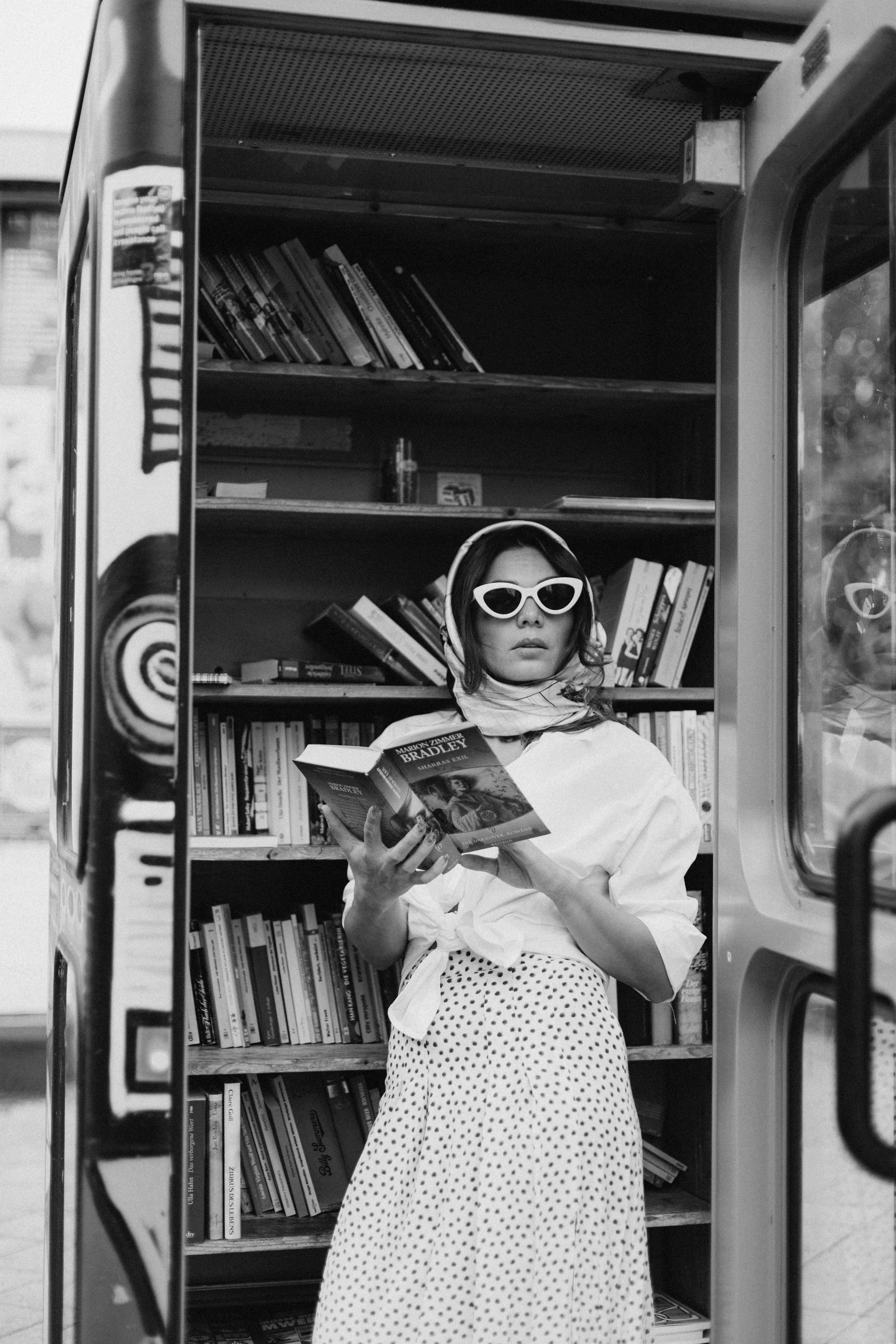 Black and white photo of a stylish woman reading a book in an outdoor library kiosk.