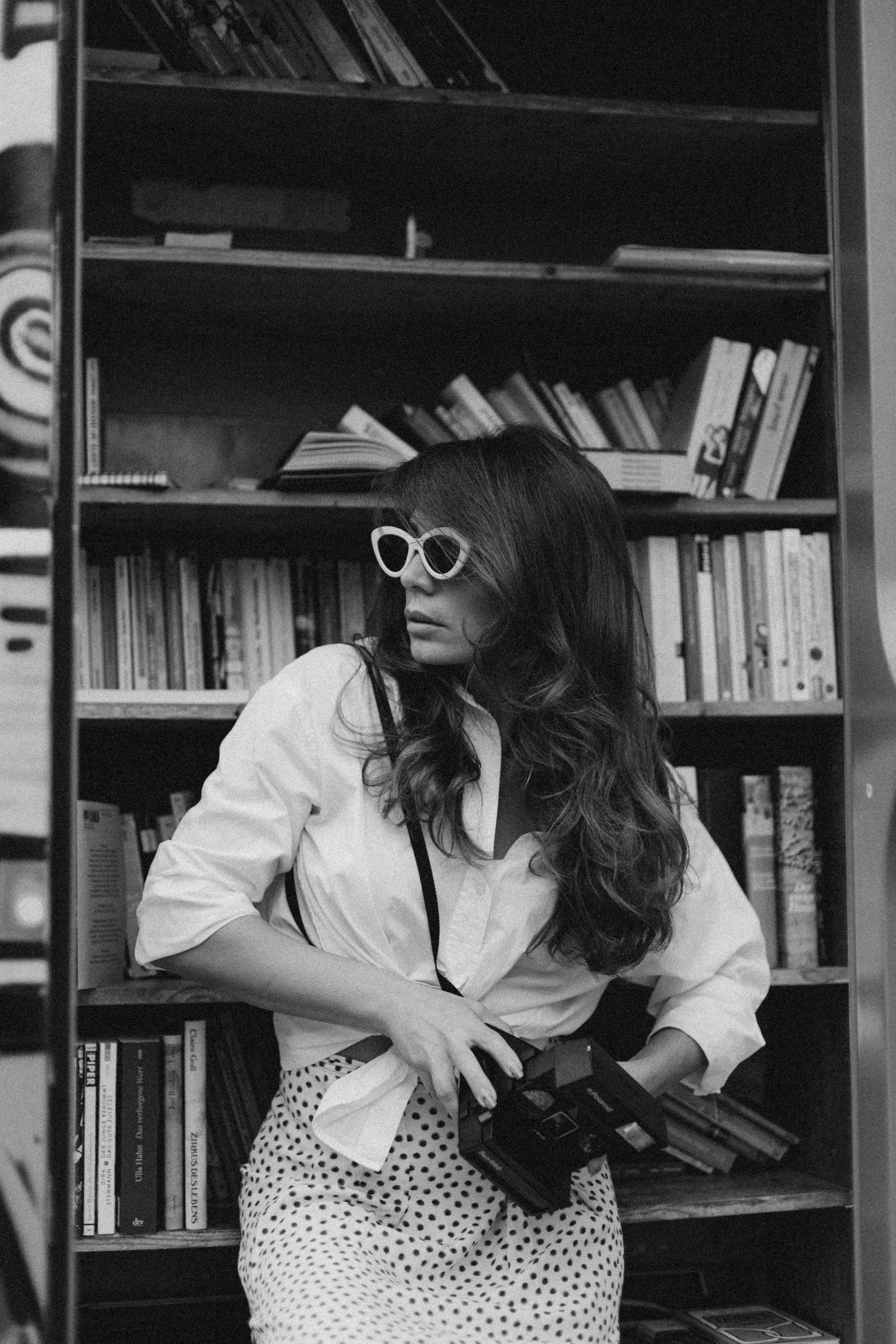 Black and white portrait of a stylish woman holding a camera in a vintage bookstore setting.