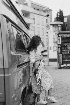 A woman in casual attire leans on a classic van in an urban setting, evoking nostalgia.