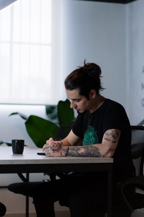 Free Tattooed woman writing at desk in minimalistic office with a coffee cup. Stock Photo